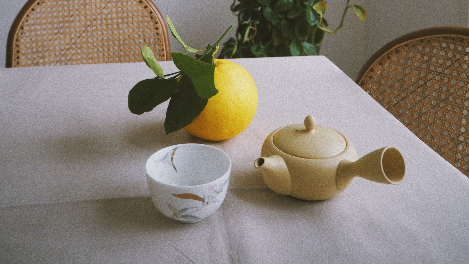 Tea set with a teapot and cups on a table with a plant in the background.